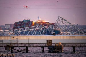 A TWISTED BRIDGE STRUCTURE LIES ABOVE A SEA CHANNEL, A HUGE CONTAINER SHIP IN THE BACKGROUND.