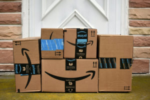 SEVERAL DIFFERENT-SIZED AMAZON BOXES SIT IN FRONT OF A WHITE DOOR SURROUNDED BY BRICKS.