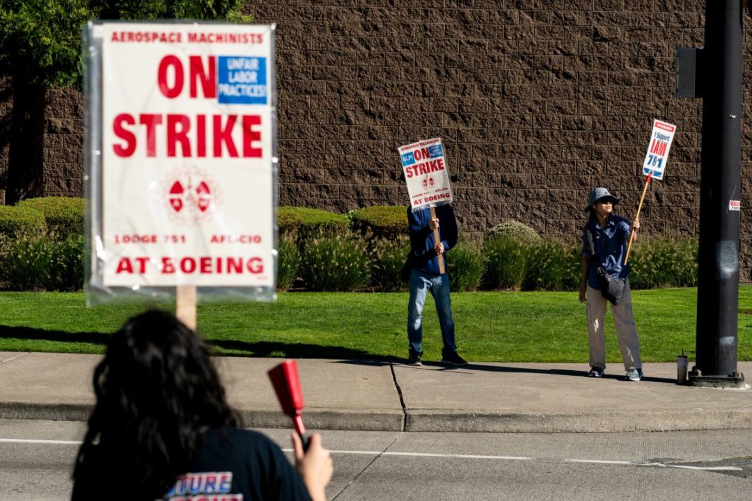 A WOMAN HOLDING A SIGN SAYING ON STRIKE AT BOEING AND A RED COWBELL FACES OTHER STRIKE WORKERS ACROSS A STREET HOLDING SIMILAR SIGNS