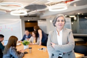 A GREY-HAIRED WOMAN IN A SHARP SUIT STANDS SMILING AT THE END OF A CONFERENCE TABLE WHERE OTHERS ARE GATHERED