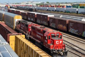 A red Canadian Pacific train in the foreground pulling yellow railcars, with a row of brown railcars on an adjacent track