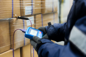 A WORKER WEARING A JACKET AND GLOVES IS HOLDING A THERMOMETER TO READ THE TEMPERATURE OF BOXES.