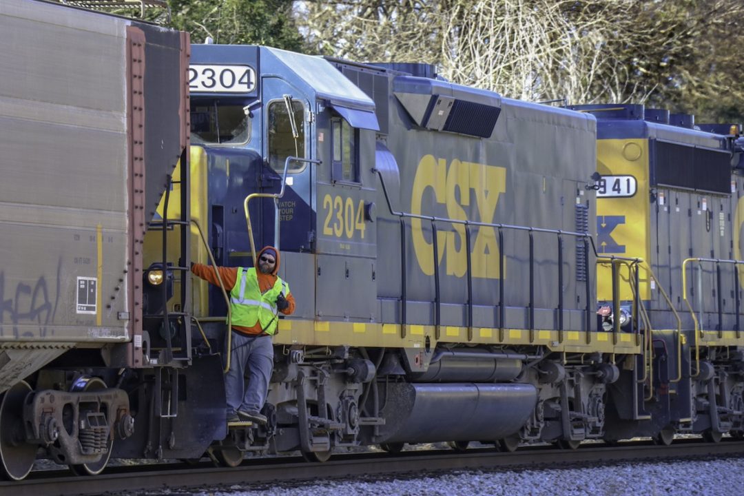 A blue train engine with yellow writing along the side that reads "CSX," with a worker in a yellow vest leaning out the side