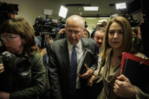 A BALDING, GREY-HAIRED MAN IN A SUIT IS USHERED THROUGH A CLAMORING CROWD OF REPORTERS WITH CAMERAS IN HAND BY A WOMAN HOLDING A CLIPBOARD