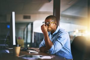  A MAN AT AN OPEN LAPTOP PINCHES THE BRIDGE OF HIS NOSE IN FRUSTRATION