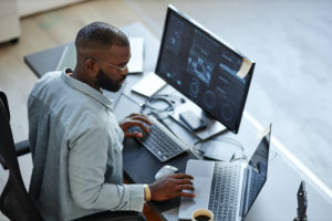 A PERSON SITS AT A DESK IN FRONT OF A LAPTOP AND A DESKTOP COMPUTER WITH DATA ON BOTH SCREENS. THE PERSON IS LOOKING TO THEIR RIGHT AT THE LAPTOP WITH THEIR HANDS ON BOTH KEYBOARDS.