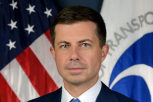 A HANDSOME MAN IN A SUIT AND TIE STANDS IN FRONT OF AN AMERICAN FLAG