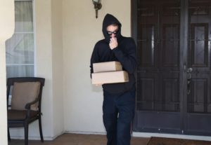 A man in a black hoodie pulled over his face, holding two packages on a front porch