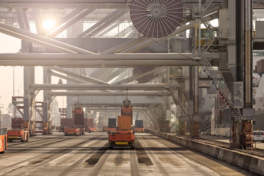 A row of white shipping cranes loading containers onto platforms as the sun sets in the background