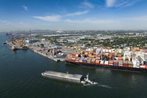 A large blue container ship docked at a port, below three white shipping cranes, while a grey barge moves through the waterway in the foreground