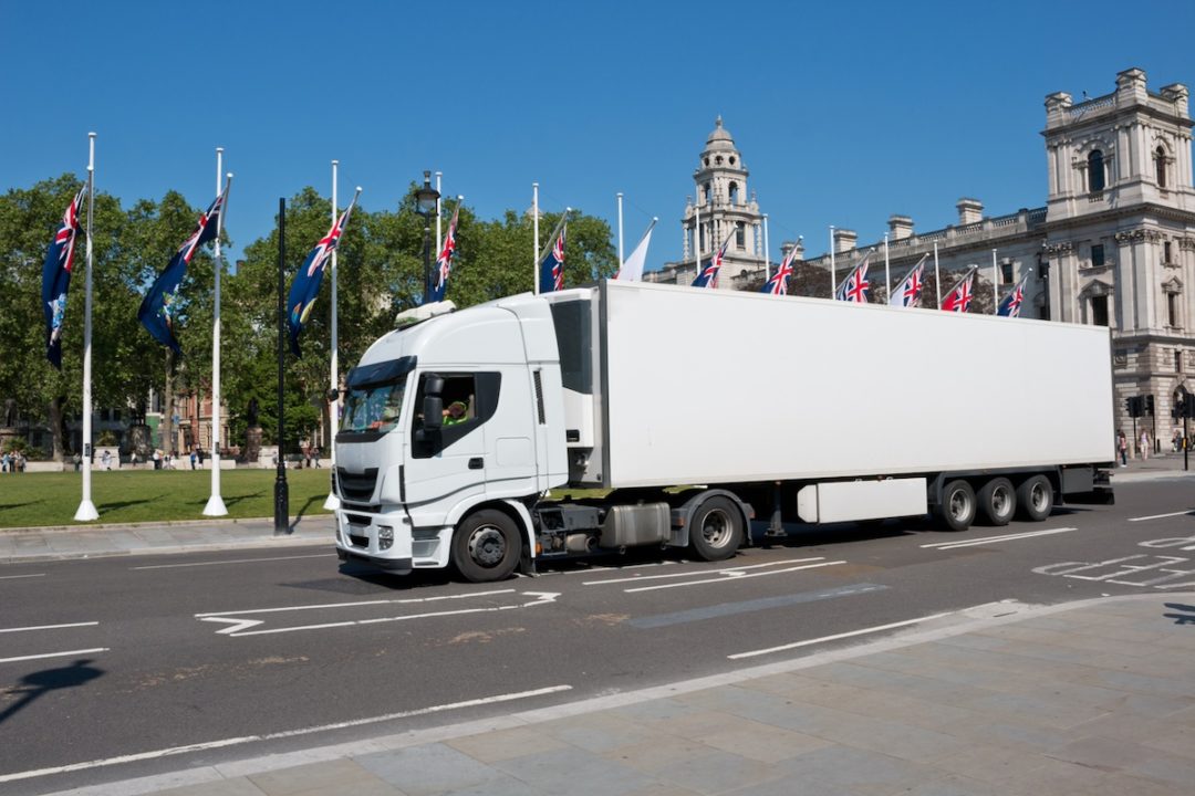 A white semi-truck pulling a white cab, along a road lined with British flags. 