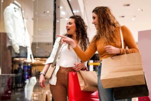 TWO WOMEN, HOLDING SHOPPING BAGS, LAUGH AND GESTURE AT AN INDOOR STOREFRONT.