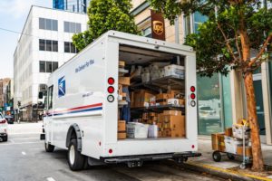 A white USPS delivery truck with the back door opened, with stacks of packages and mail inside