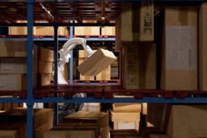 A robotic arm holding a package in front of a blue shelf stacked with other boxes