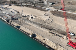 Cranes and construction workers on a bare gravel plot of land alongside a waterway